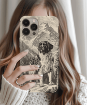 Beige and black phone case featuring a detailed vintage-style illustration of a Saint Bernard standing proudly against a snowy mountain landscape. Classic alpine dog design with rustic charm, perfect for nature and dog lovers.