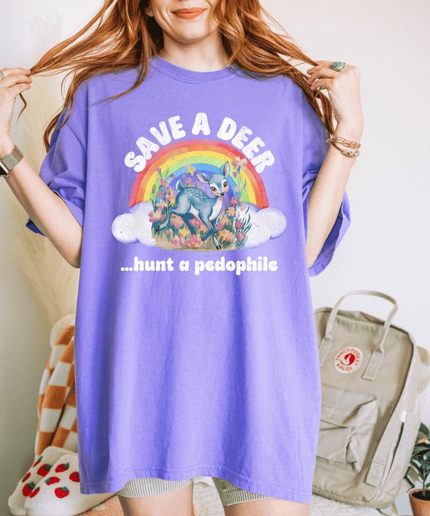 A violet purple t-shirt featuring a cute illustrated deer under a rainbow with clouds and flowers, surrounded by the bold, humorous text “Save a Deer… Hunt a Pedophile” in vintage distressed white font.