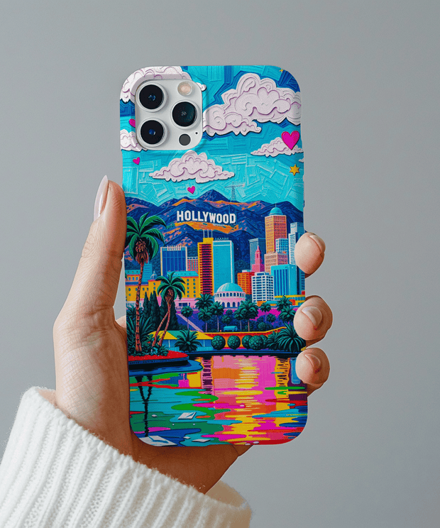 A vibrant phone case showcasing a colorful Hollywood skyline with palm trees, skyscrapers, mountains, pink hearts, and a glowing sunset reflection in rainbow tones. The design radiates retro California vibes.