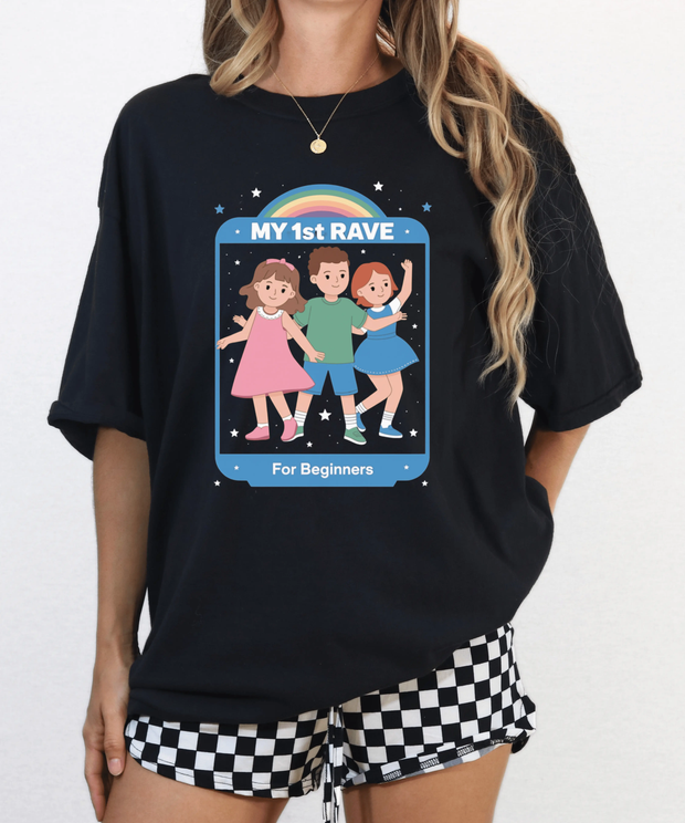A black oversized t-shirt featuring a retro cartoon-style print of three dancing kids under a rainbow with stars around them. The design reads “MY 1st RAVE – For Beginners,” creating a playful, nostalgic, and ironic party vibe.