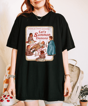 A black oversized t-shirt featuring a retro children’s book-style illustration of kids gathered around a summoning circle with the phrase “Let’s Summon Demons” in vintage red text, parodying wholesome 80s design aesthetics.