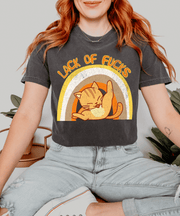 A gray oversized t-shirt featuring a retro-style orange cat stretching inside a warm-toned rainbow graphic with the phrase “Lack of F*cks” written above in distressed vintage lettering.