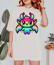 Ivory oversized t-shirt featuring a cute kawaii Baphomet character with rainbow wings, striped rainbow body, colorful horns, and sparkly stars. Bright, pastel goth design mixing adorable chaos and pride energy.