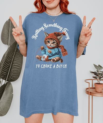 A blue jean colored oversized t-shirt featuring a cute kitten wearing a red hat and scarf, knitting yarn, with the funny text “Knitting Something Fancy To Choke A Bitch” in bold white vintage lettering.