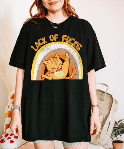 A black oversized t-shirt featuring a retro-style orange cat stretching inside a warm-toned rainbow graphic with the phrase “Lack of F*cks” written above in distressed vintage lettering.