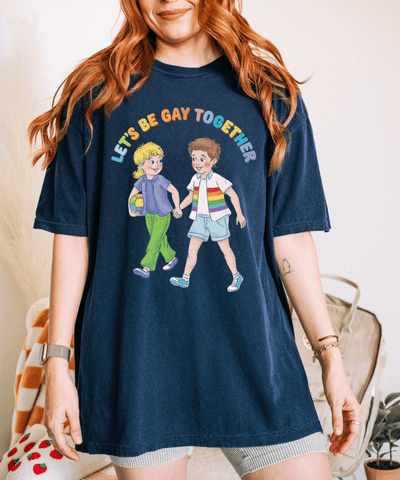A navy blue oversized t-shirt with a vintage-style illustration of two kids holding hands, one wearing a rainbow-striped shirt, and the phrase “Let’s Be Gay Together” in colorful retro lettering above them.