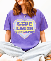 A woman wearing a purple oversized t-shirt with a retro pastel heart graphic that reads “Live Laugh Lorazepam” in 70s bubble letters. The design features soft yellow, lavender, and pink colors for an ironic vintage aesthetic.