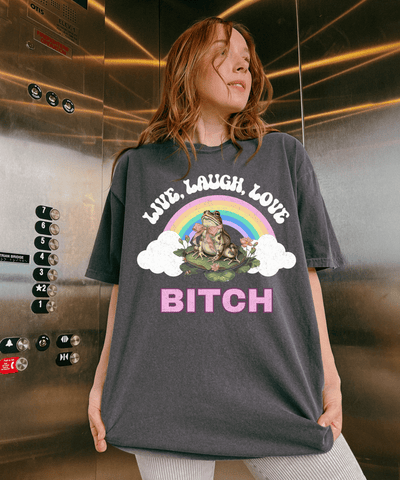 A woman wearing a gray oversized t-shirt with a colorful rainbow graphic and a frog sitting on a lily pad. The shirt says “Live Laugh Love Bitch” in bold retro-style letters for a sassy, ironic aesthetic.