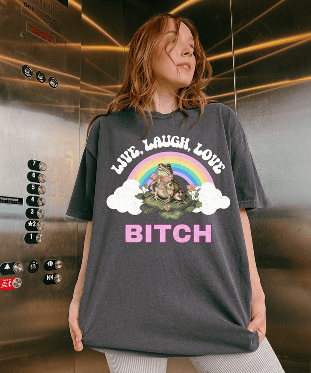 A woman wearing a gray oversized t-shirt with a colorful rainbow graphic and a frog sitting on a lily pad. The shirt says “Live Laugh Love Bitch” in bold retro-style letters for a sassy, ironic aesthetic.
