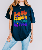 A navy oversized t-shirt with bold rainbow retro bubble letters reading “LOUD PROUD FEMINIST AS F***,” accented by colorful hearts. The design radiates empowerment and 70s-inspired pride energy.
