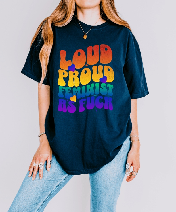 A navy oversized t-shirt with bold rainbow retro bubble letters reading “LOUD PROUD FEMINIST AS F***,” accented by colorful hearts. The design radiates empowerment and 70s-inspired pride energy.