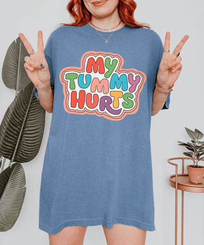Blue oversized t-shirt with a bold, colorful bubble letter design that says “My Tummy Hurts.” The playful text features red, blue, green, pink, yellow, and purple letters outlined with a soft pastel border for a fun, sarcastic vibe.