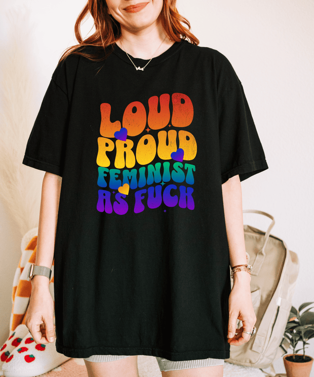 A black oversized t-shirt with bold rainbow retro bubble letters reading “LOUD PROUD FEMINIST AS F***,” accented by colorful hearts. The design radiates empowerment and 70s-inspired pride energy.