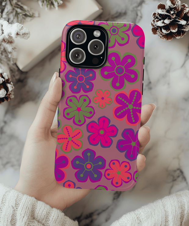 Phone case with a 70s-inspired retro flower pattern featuring bold pink, purple, green, and orange daisy shapes on a mauve background. The colorful floral design has a playful, groovy aesthetic perfect for vintage lovers.
