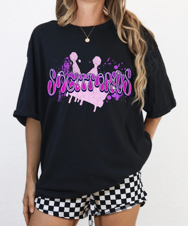 Black oversized t-shirt with the word “Sagittarius” in bold pink and purple airbrush-style bubble letters over a paint splatter background. Perfect for astrology lovers and Sagittarius signs who love edgy, vibrant, streetwear-inspired designs.