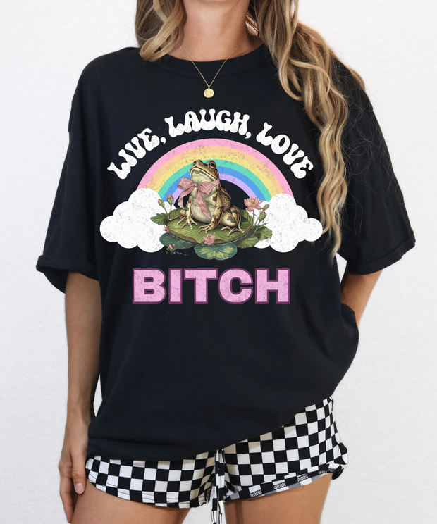 A woman wearing a black oversized t-shirt with a colorful rainbow graphic and a frog sitting on a lily pad. The shirt says “Live Laugh Love Bitch” in bold retro-style letters for a sassy, ironic aesthetic.