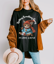 A black oversized t-shirt featuring a cute kitten wearing a red hat and scarf, knitting yarn, with the funny text “Knitting Something Fancy To Choke A Bitch” in bold white vintage lettering.