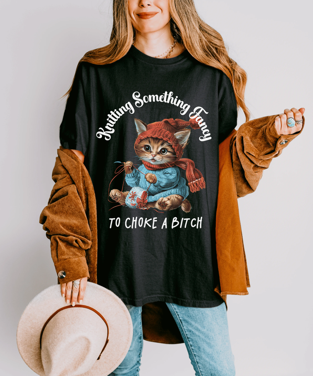 A black oversized t-shirt featuring a cute kitten wearing a red hat and scarf, knitting yarn, with the funny text “Knitting Something Fancy To Choke A Bitch” in bold white vintage lettering.