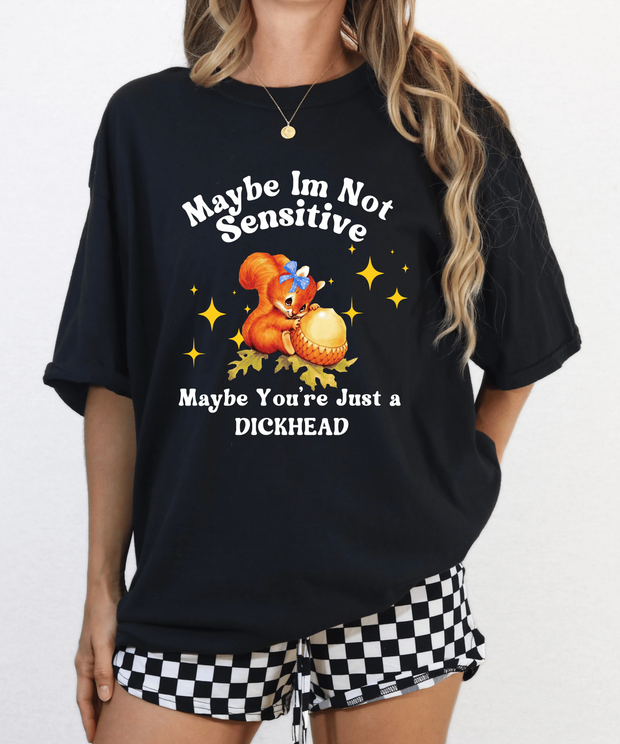 A black oversized t-shirt featuring a vintage-style cartoon squirrel holding a giant acorn with sparkles around it. The shirt reads “Maybe I’m Not Sensitive, Maybe You’re Just a Dickhead” in playful white text.