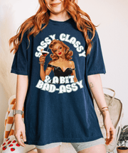 Vintage-inspired navy blue t-shirt featuring a glamorous retro pin-up woman in a black dress holding a cocktail. The text reads “Sassy Classy & A Bit Bad-Assy” in bold lettering, perfect for confident, fun-loving women with attitude.
