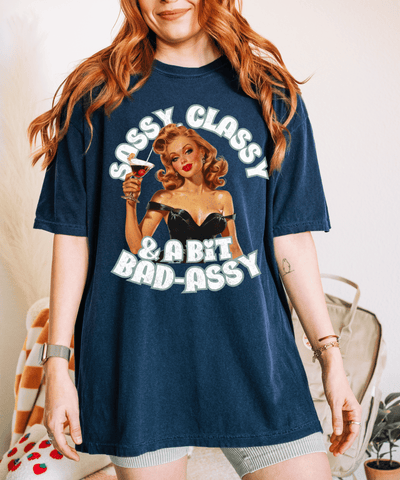Vintage-inspired navy blue t-shirt featuring a glamorous retro pin-up woman in a black dress holding a cocktail. The text reads “Sassy Classy & A Bit Bad-Assy” in bold lettering, perfect for confident, fun-loving women with attitude.