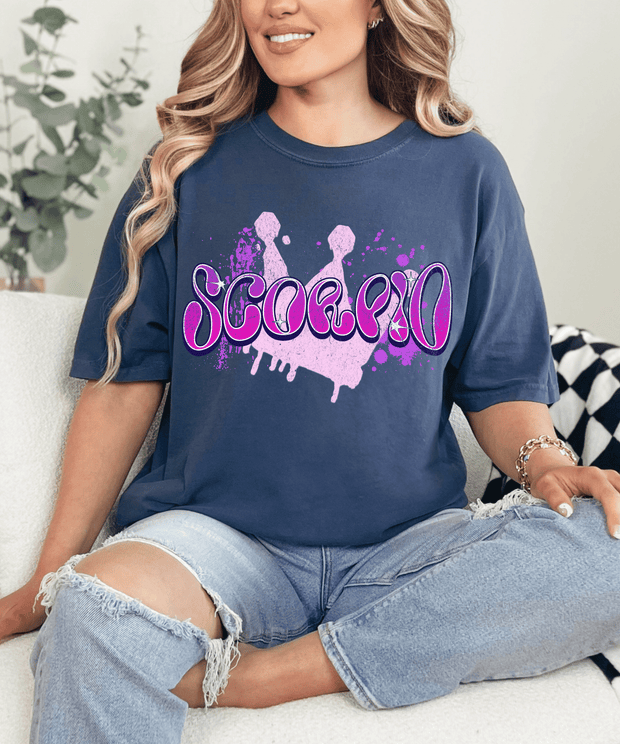 Navy blue oversized t-shirt with bold purple and pink airbrush-style lettering that spells “Scorpio.” The design features paint splatters and a grunge background, perfect for astrology lovers and Scorpio zodiac fans who love edgy, retro streetwear style.