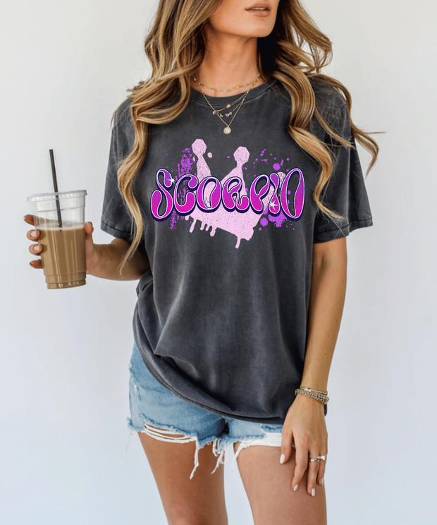 Pepper gray oversized t-shirt with bold purple and pink airbrush-style lettering that spells “Scorpio.” The design features paint splatters and a grunge background, perfect for astrology lovers and Scorpio zodiac fans who love edgy, retro streetwear style.