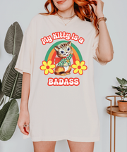 Ivory oversized t-shirt with a vintage-style graphic of a cute kitten wearing a plaid scarf and red cape. Surrounded by a retro rainbow arch and yellow flowers, the text reads “My Kitty Is a Badass” in bold red bubble letters.
