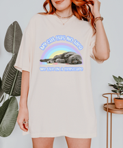 An ivory oversized t-shirt featuring a gray cat lying lazily under a pastel rainbow with the text “My Cat Has No Idea We Live in a Hellscape.” The design blends cute cat imagery with dark humor in a soft retro aesthetic.