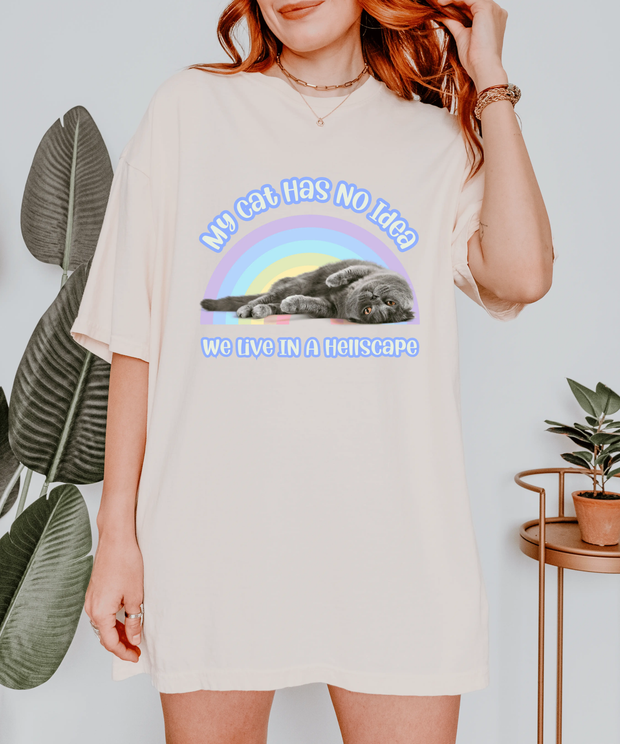 An ivory oversized t-shirt featuring a gray cat lying lazily under a pastel rainbow with the text “My Cat Has No Idea We Live in a Hellscape.” The design blends cute cat imagery with dark humor in a soft retro aesthetic.