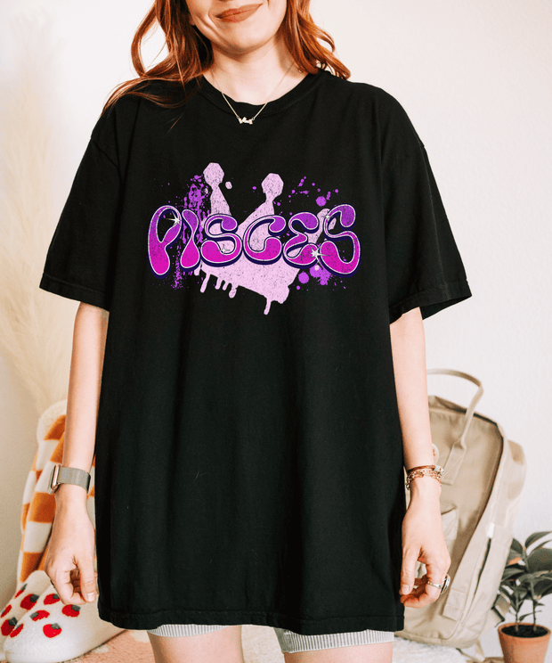 Black oversized t-shirt with the word “Pisces” in a purple graffiti-style drip font and ink-splatter accents. The design gives off a bold, artsy zodiac vibe perfect for astrology lovers and those with a creative, dreamy edge.